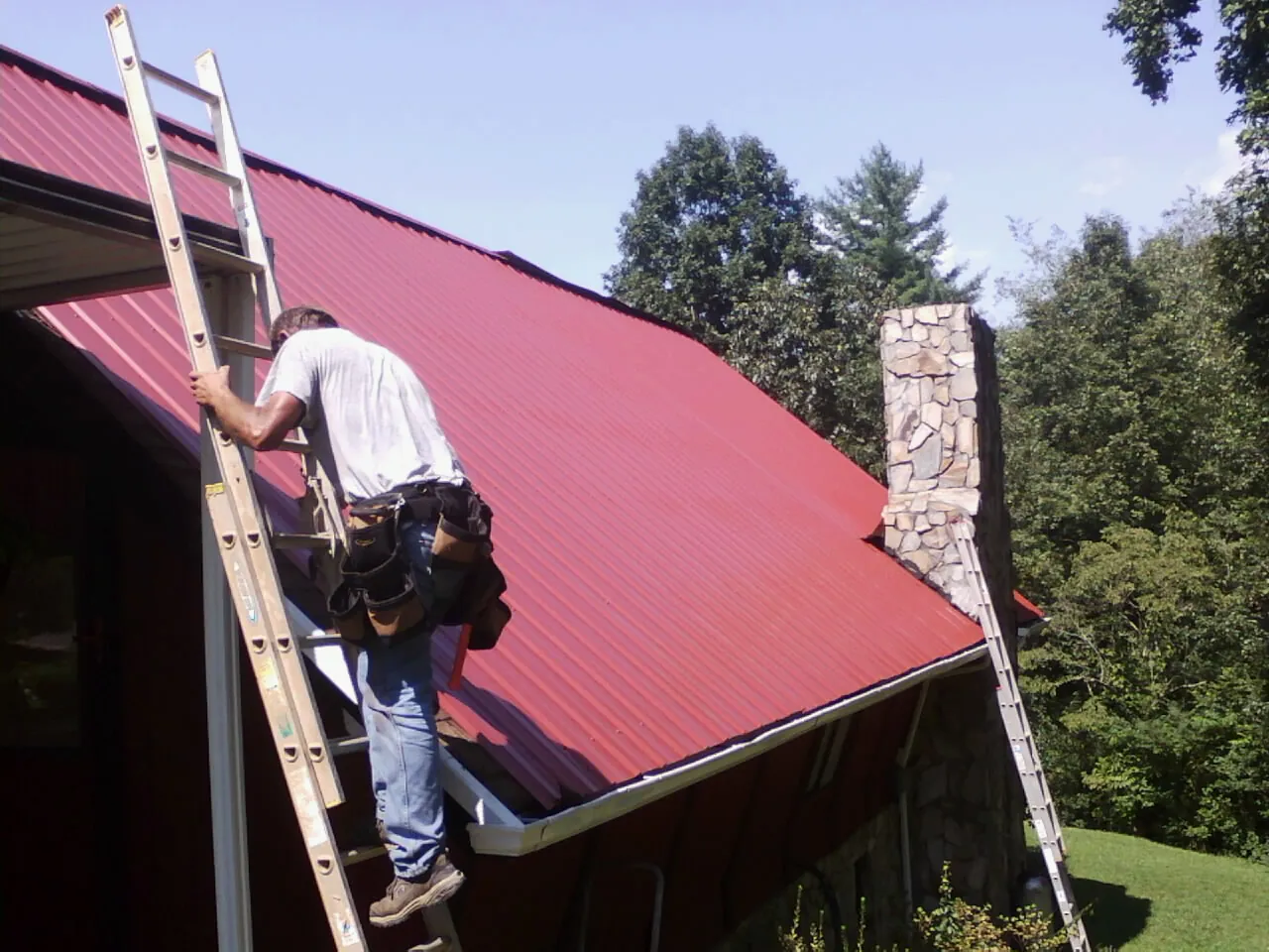 Roofer on a ladder beside a red metal roof and stone chimney — Soffit & Fascia Repair by Roofing Brothers in Sebago