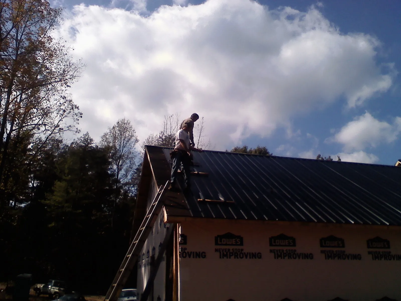 Roofer fastening dark metal panels high against a cloudy sky, from a Roof Inspection job in Sebago